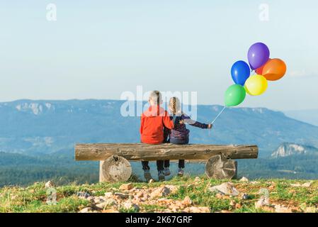 Une fille et un garçon sont assis sur un banc avec des ballons balançant dans le vent. Vue de l'arrière. Montagne en arrière-plan Banque D'Images