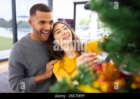 Image de heureux couple divers célébrant noël à la maison décorant l'arbre Banque D'Images