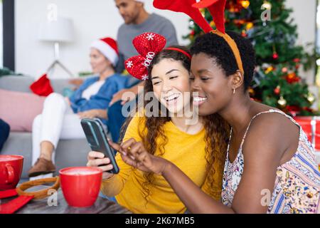 Image d'amis féminins heureux et variés utilisant un smartphone, célébrant noël avec des amis variés Banque D'Images