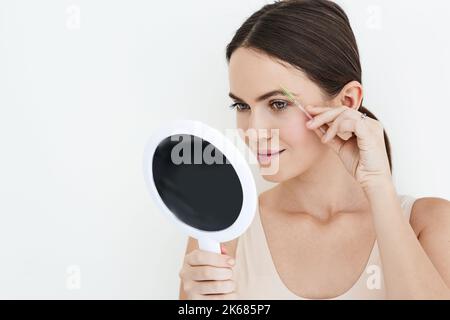 Belle femme peignant ses sourcils avec un outil de brosse tout en regardant dans le miroir portable, isolé sur blanc. Processus de brossage des sourcils Banque D'Images