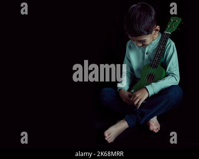 latino garçon assis pieds nus sur le sol avec un ukulele vert dans ses bras riant et regardant vers le bas. Photo studio avec arrière-plan noir et espace de copie Banque D'Images