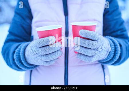 Une femme en gants tient dans ses mains deux tasses en papier rouge de boisson chaude en hiver sur la nature, un thé chaud en saison froide, une promenade et un petit voyage en hiver. Photo de haute qualité Banque D'Images