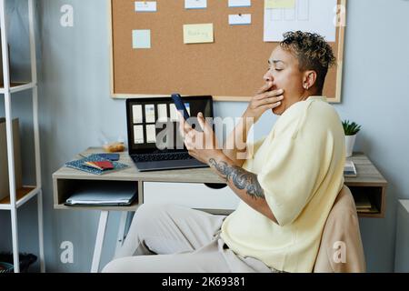 Portrait en vue latérale d'une femme noire tatouée bâillant sur le lieu de travail du bureau et défilant sur les médias sociaux par téléphone Banque D'Images