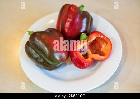 Trois poivrons rouges cultivés à la maison, qui se transforment juste en rouge avec une coupe en moitié sur une assiette blanche. ÉTATS-UNIS. Banque D'Images