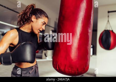 Déterminé boxer femelle poinçonné un sac rouge pendant l'entraînement à la salle de gym. Jeune femme athlétique pratiquant ses techniques de poinçonnage dans un gymnase de boxe. Banque D'Images