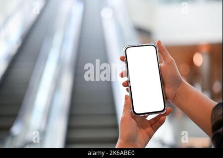 Une maquette d'écran blanc de smartphone se trouve entre les mains d'une femme sur l'escalier mécanique du centre commercial flou en arrière-plan. Gros plan Banque D'Images