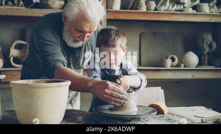 Un petit garçon concentré apprend à travailler avec de l'argile sur une roue de jet professionnelle en cours de poterie dans un atelier traditionnel. Son professeur senior expérimenté l'aide. Banque D'Images