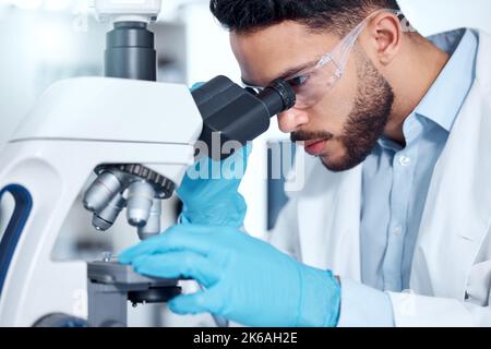 Un beau jeune homme de course mixte avec une barbe sombre portant des gants et un manteau et regardant des échantillons médicaux au microscope dans son laboratoire. Un homme Banque D'Images