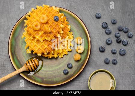 Un petit-déjeuner en bois, des baies et des gaufres sur une assiette verte. Bol vert avec miel et bleuets sur la table. Pose à plat. Arrière-plan gris. Banque D'Images