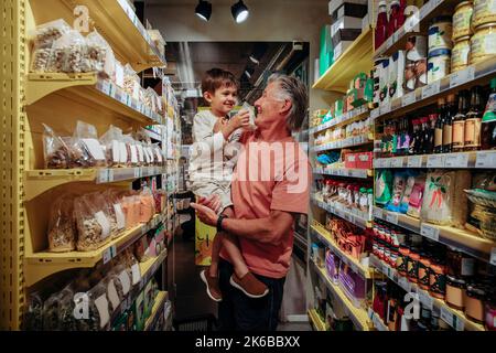 Homme senior souriant transportant son petit-fils tout en faisant du shopping dans un supermarché Banque D'Images