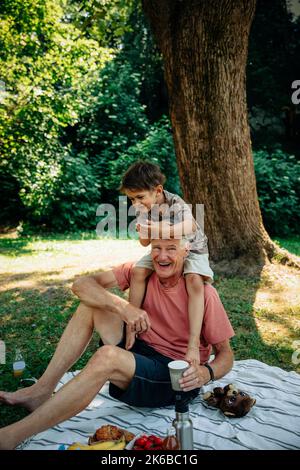 Grand-père portant le petit-fils sur l'épaule tout en étant assis sur une couverture de pique-nique au parc Banque D'Images