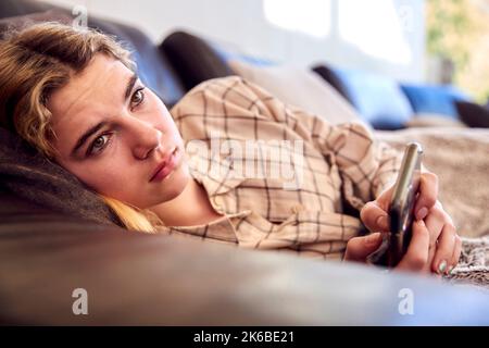 Adolescente malheureuse mentant sur un canapé avec téléphone mobile victime d'intimidation en ligne à la maison Banque D'Images