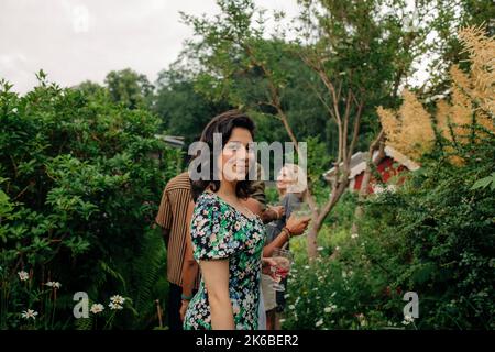 Vue latérale d'une femme souriante avec boisson debout près des plantes dans le jardin Banque D'Images