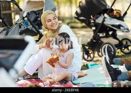 Femme souriante dans le hijab regardant loin tout en étant assise avec la fille tenant l'ours en peluche Banque D'Images