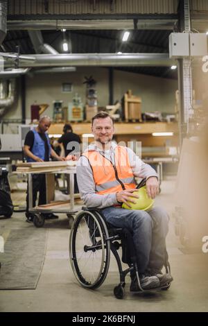 Portrait d'un charpentier souriant assis en fauteuil roulant avec des collègues travaillant en arrière-plan à l'entrepôt Banque D'Images