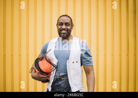 Portrait d'un ouvrier de construction homme confiant avec un casque de sécurité debout contre un mur en métal jaune Banque D'Images