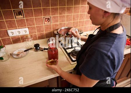 Vue arrière d'une femme de ménage qui verse du jus de tomate fraîchement bouilli dans un pot stérilisé, préparant des passata biologiques faits maison Banque D'Images