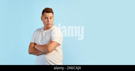 Portrait d'un jeune garçon, modèle en surpoids en chemise blanche debout avec bras croisés isolés sur fond bleu. Concept de la jeunesse, des émotions, du visage Banque D'Images