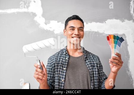 Un jeune homme asiatique tient des échantillons de peinture et des rouleaux sur le point de peindre un mur réparé Banque D'Images