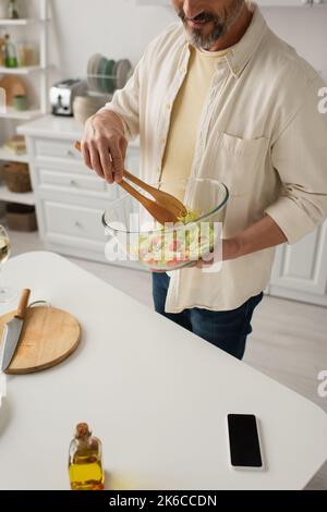 vue partielle de l'homme souriant mélangeant de la salade de légumes frais avec des pinces en bois tout en cuisinant dans la cuisine, image de stock Banque D'Images