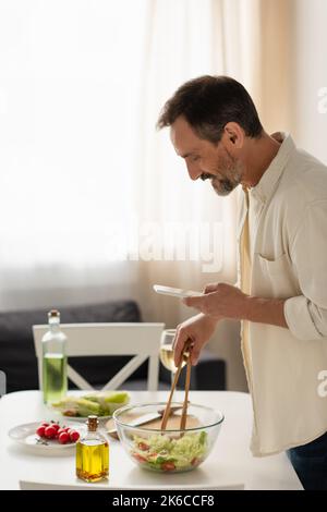 vue latérale de happy man mélange de salade de légumes avec des pinces en bois tout en regardant la recette dans le téléphone mobile, image de stock Banque D'Images