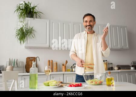 homme heureux avec un smartphone et des pinces en bois regardant l'appareil photo près des légumes frais et des bouteilles avec de l'huile, image de stock Banque D'Images