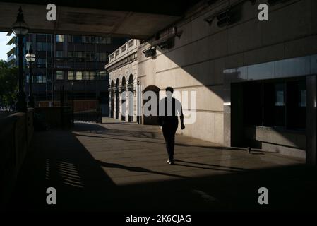 Un homme marche en silhouette sur le quai du London Bridge sous le London Bridge pris dans la lumière du soleil du soir. Banque D'Images