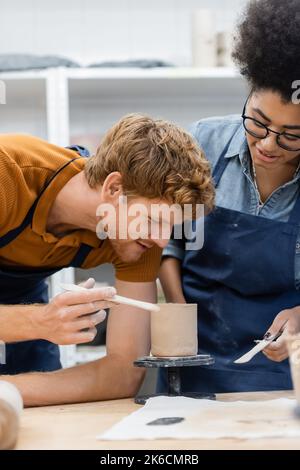 Souriant redhead enseignant tenant l'outil près de coupe d'argile et femme afro-américaine dans l'atelier de poterie, image de stock Banque D'Images