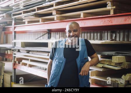 Portrait du charpentier chauve souriant debout par pile de palettes dans l'entrepôt Banque D'Images