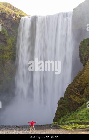 L'homme avec son bras en l'air debout au pied de la cascade de Skogafoss, Islande Banque D'Images