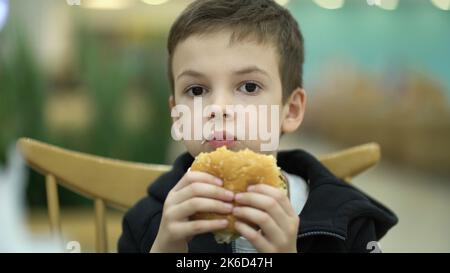 Un garçon triste et fatigué de sept ans mange un hamburger dans une aire de restauration après une journée d'école. Enfant dans un sweat-shirt en dégustant un hamburger juteux Banque D'Images