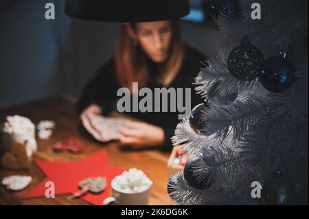 Vue rapprochée d'un arbre de noël blanc décoré de boules de Noël bleu brillant placées sur une table domestique avec une jeune femme floue emballant des cadeaux de vacances Banque D'Images