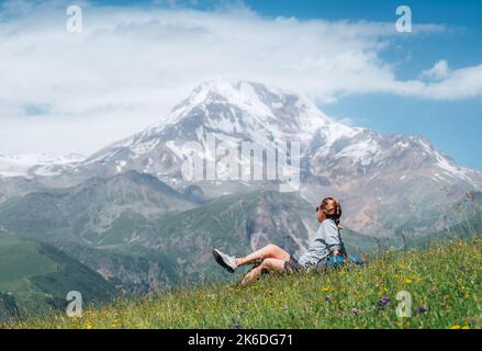 Backpacker femme assise sur une colline d'herbe verte et appréciant les pentes enneigées de la montagne Kazbek 5054m avec un sac à dos pendant qu'elle marche par l'herbe verte hil Banque D'Images