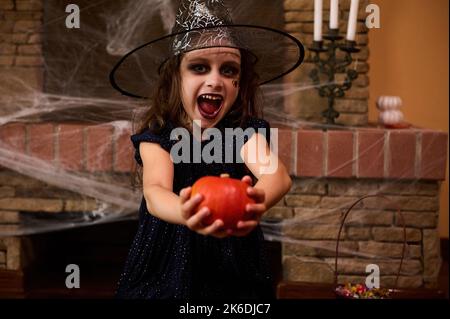 Charmante petite fille, vêtue de sorcière, riant et donnant une citrouille à la caméra, assise contre la cheminée avec toile d'araignée Banque D'Images