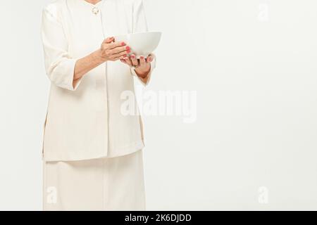 La vieille femme porte des vêtements blancs classiques tenant un bol vide isolé sur fond blanc Banque D'Images