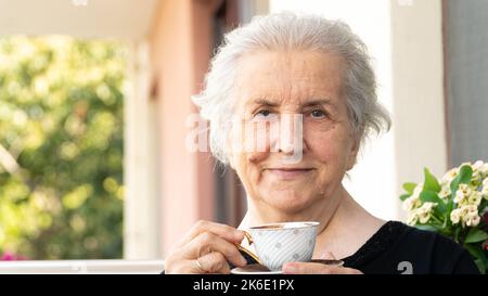 Vieille femme souriante regardant un appareil photo, buvant un café turc et se détendant sur le balcon. Femme âgée insouciante en 70s souriant et appréciant le temps. Banque D'Images