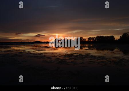 Photo panoramique d'un coucher de soleil dans des tons orange et bleu avec le reflet du ciel sur l'eau et des arbres ombrages le long de celle-ci Banque D'Images