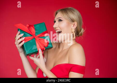 Surprise, femme blonde souriante riant dans la robe de gala à col bas, recevoir boîte cadeau dans le studio rouge. Offre de remise Banque D'Images