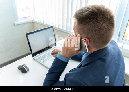 Homme d'affaires concentré ayant un appel téléphonique et travaillant sur son ordinateur portable dans son bureau. Un directeur dans un bureau lumineux habillé d'un bleu costume tal Banque D'Images