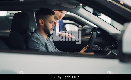 L'acheteur de voiture homme confiant est assis dans le siège du conducteur dans la nouvelle automobile dans la salle d'exposition moderne et parle au vendeur debout près de la fenêtre de voiture. Guy tient le volant. Banque D'Images