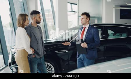 Un jeune couple intéressé, homme et femme, parle au directeur de la concession automobile pendant qu'il leur parle du modèle automobile de luxe. Personnes magnifiques, transport et concept d'affaires. Banque D'Images