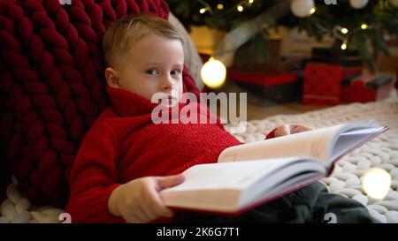 Le garçon lit un livre sur le fond de l'arbre de Noël. Gros plan du livre de lecture pour Garçon avec arbre de Noël illuminé en arrière-plan. Banque D'Images