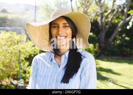 Portrait d'une femme biraciale heureuse en chapeau de soleil debout dans le jardin ensoleillé souriant Banque D'Images