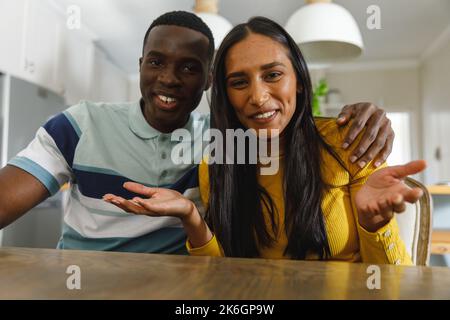 Joyeux couple divers faisant des appels vidéo souriant et faisant des gestes à la caméra dans la cuisine Banque D'Images