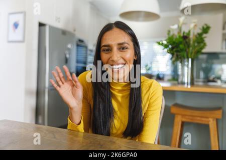 Bonne femme biraciale faisant un appel vidéo souriant et en agitant à la caméra dans la cuisine Banque D'Images