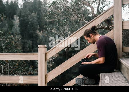 homme caucasien avec de longs cheveux recueillis avec la barbe et le t-shirt assis calme et détendu sur les escaliers du point de vue à côté de la main courante en bois Banque D'Images