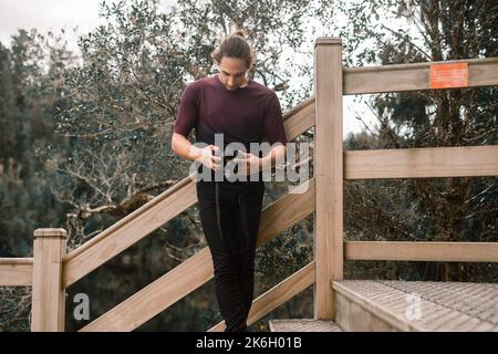 homme caucasien avec de longs cheveux recueillis avec le t-shirt ajusté à la barbe et le pantalon noir debout à pieds croisés regardant l'écran de l'appareil photo calme et détendu sur Banque D'Images