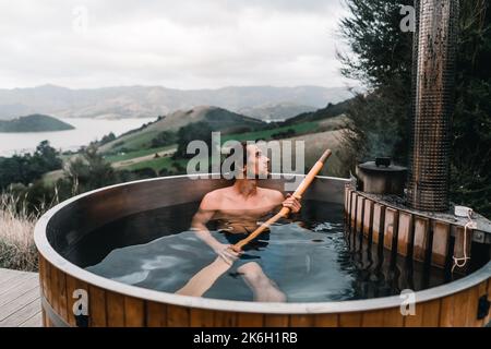 calme garçon caucasien sans vêtements prenant un bain avec une longue cosse en bois dans ses mains à l'intérieur d'un jacuzzi d'eau chaude métallique se relaxant dans les montagnes Banque D'Images