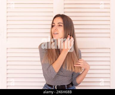 Jeune belle femme blonde heureusement se touchant et se brossant les cheveux à la main tout en regardant sur le côté dans fond blanc de porte Banque D'Images