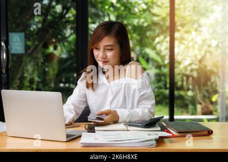 Jeune femme asiatique en tenue élégante et décontractée tenant un smartphone et travaillant sur un ordinateur portable tout en étant assise près de la fenêtre dans le bureau à domicile. Banque D'Images